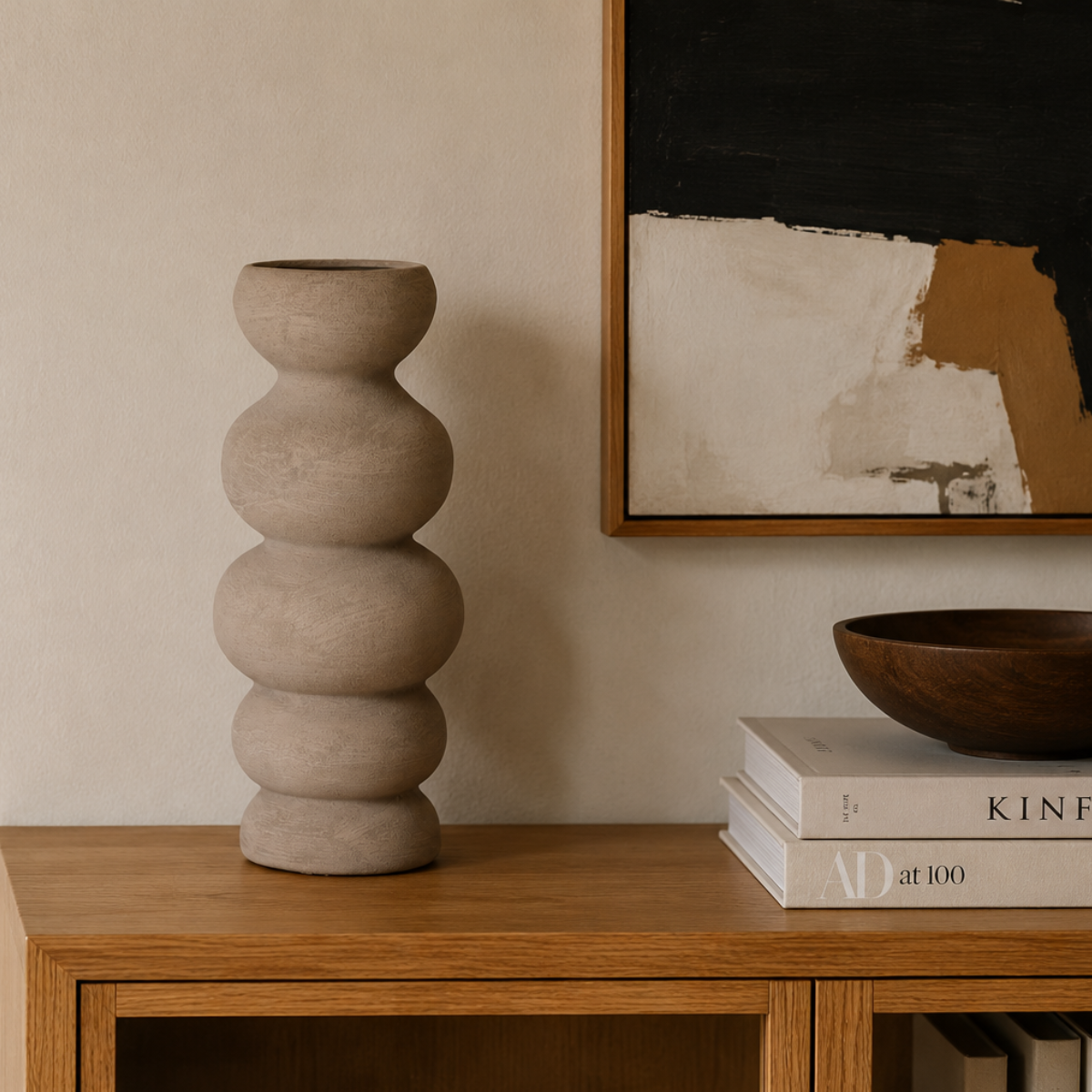 Jarrón decorativo beige de formas redondeadas sobre mueble de madera junto a cuadro abstracto