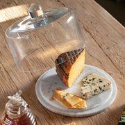 Quesera con base de mármol blanco y campana transparente con diferentes quesos sobre una mesa de madera.
