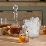 Licorera y cubitera de vidrio tallado con hielo, acompañadas de vasos sobre una mesa.